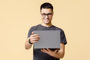 Online education and work, message from social networks and good offer. Cheerful young man student with glasses looks at laptop and reads chat, isolated on beige background, studio shot photo