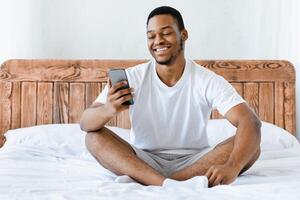 Positive Black Guy Using Cellphone Making Call Online Sitting Using Phone In Bed At Home In Modern Bedroom. Distant Communication, Mobile Conference Application Concept photo