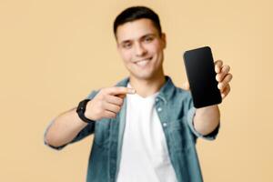 It is cool, best mobile app and digital ad. Happy young man in casual shows finger at smartphone with blank screen, selective focus on device, isolated on beige background, studio shot, free space photo