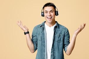 Relax, favorite music, fun and pleasure at spare time. Smiling excited student man in casual with modern headphones spreads his hands in sides, isolated on light sandy background, empty space photo