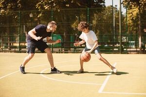 Full body length portrait in action of cheerful man and boy playing basketball on a sports court, little curly guy leading the ball and dribbling, excited man teaching, spending free time together photo