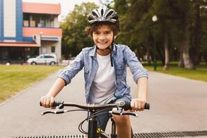 Summer Activity. Front portrait of cheerful little boy riding a bicycle in public park and smiling, wearing protective safety helmet, posing and looking at camera, blurred background, selective focus photo