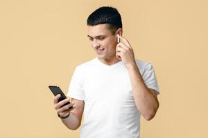 Favorite playlist for walking and sports. Smiling guy in t-shirt in modern wireless headphones looks in smartphone and turns on playlist, isolated on beige background, studio shot, free space photo