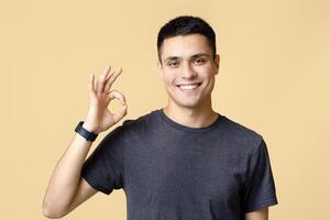 Great job, good mood and cool review. Smiling young attractive guy student with smart watch shows hand gesture ok and look at camera, isolated on beige background, studio shot, free space photo
