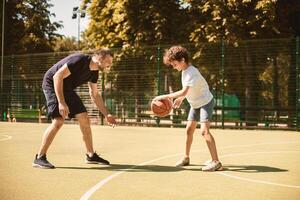 Sports Concept. Side view portrait in action of man and boy playing basketball together on the field photo