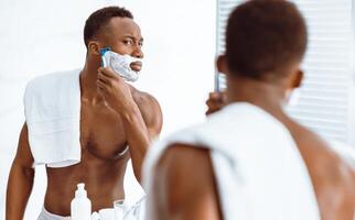 Shirtless Black Guy Shaving Face In The Morning Using Shave Foam And Razor Looking In Mirror Standing In Modern Bathroom At Home. Facial Skin Care Products Advertisement. Selective Focus photo