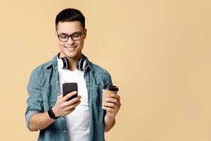 Modern worker communication in social networks and typing message. Smiling attractive young guy in glasses, casual, with headphones texting on phone and hold coffee cup, isolated on beige background photo