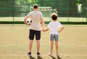 Sports Concept. Back view of dad and son standing at football pitch and looking at goal net, holding hands photo