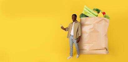 A customer stands holding a smartphone next to a large paper bag filled with fresh groceries. The vibrant background enhances the shopping experience in a modern market setting. photo