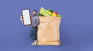 A person stands next to a paper grocery bag filled with fresh produce while holding a smartphone. This represents the shift in shopping behavior in today's isolated environment. photo