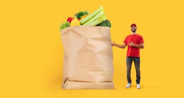 A delivery man stands proudly next to a large paper bag filled with fresh fruits and vegetables, reflecting the new normal of grocery shopping amidst isolation, in a vibrant store setting. photo
