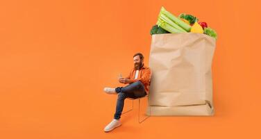 A person sits on a chair with a smartphone in hand, looking engaged as they shop for groceries. Beside them is a large paper bag filled with colorful vegetables, indicating a focus on healthy eating. photo