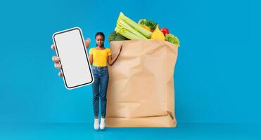 A customer stands next to a large paper bag filled with fresh groceries while holding a smartphone. This reflects modern shopping trends, emphasizing convenience and delivery services. photo