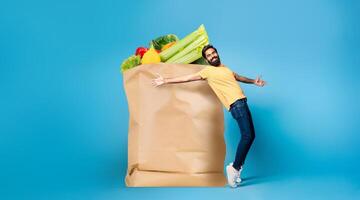 A man in casual attire appears delighted while holding a large paper bag filled with fresh groceries. He stands against a blue backdrop, showcasing the joy of shopping in today's world. photo