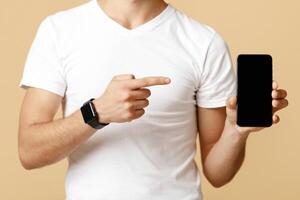 Cropped of young student man in protective mask with glasses shows finger at smartphone with blank screen, isolated on light sand background, studio shot photo