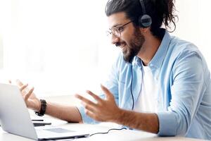 Web Conference. Friendly Arabic Freelancer Guy In Headset Making Call On Laptop, Sitting At Desk In Home Office, Talking And Gesturing At Camera, Enjoying Online Communication, Copy Space photo