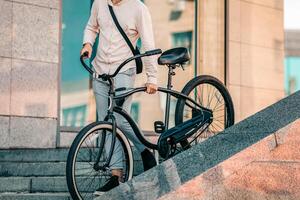 A young man carries his bicycle down a set of stairs in a city. He is dressed casually, embodying a modern lifestyle as he connects with his surroundings while admiring the urban architecture, cropped photo