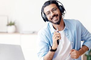 Cheerful Arab Guy In Wireless Headphones Listening Music On Smartphone And Singing While Sitting At Desk At Home Office, Joyful Eastern Man Using Cellphone Like Microphone And Smiling At Camera photo