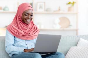 Remote Work. Smiling African Muslim Lady In Hijab Using Laptop At Home, Sitting On Couch With Computer, Enjoying Freelance Job, Working Online In Internet While Sitting On Couch With PC, Free Space photo