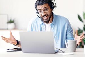 Joyful Arab Man In Headset Having Web Conference On Laptop In Office, Cheerful Eastern Guy Making Call With Computer, Sitting At Desk Talking And Gesturing At Camera, Free Space photo