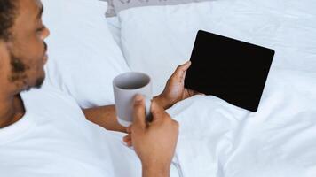 African American Man Using Digital Tablet With Empty Screen Enjoying Morning Coffee And Reading E-Book Online Lying In Bed At Home On Weekend. Internet Website Advertising. Cropped, Mockup, Panorama photo