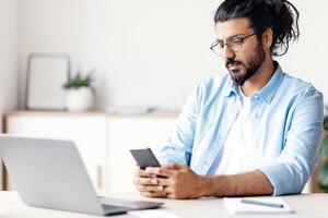 Pensive Arab Freelancer Man Messaging On Smartphone While Sitting At Desk In Home Office, Having Break In Work, Young Eastern Guy Using Cellphone, Browsing Internet Or Checking Social Networks photo