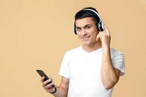 Modern gadgets and music break at home. Attractive happy zoomer blogger man in white t-shirt with modern headphones, turns on playlist on phone, isolated on light background photo