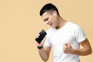 Fun, relax and joy at home in self-isolation at free time. Cheerful young man in modern wireless headphones sings in imaginary microphone at smartphone, isolated on beige background photo