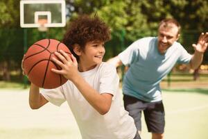 Active Leisure Concept. Portrait of cheerful father and son playing basketball together, man trying to catch the ball, boy running from him, selective focus, blurred background photo