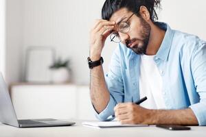 Stylish Freelancer Guy Taking Notes To Notepad While Working At Home Office, Checking His Business Timetable, Writing Plan, Sitting AT Desk With Laptop, Massaging Forehead, Copy Space photo