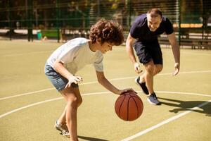 Enjoying Game. Portrait of cheerful father and son playing basketball together on a sports court, boy leading the ball and dribbling, excited man trying to take ball. Family having fun together photo