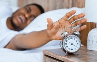 Early Awakening. Stressed African American Guy Turning Off Alarm Clock Waking Up Early In The Morning Lying In Bed At Home. Time To Wake Up Concept. Selective Focus, Shallow Depth photo