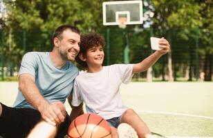 Family Selfportrait. Handsome boy taking selfie with his daddy, sitting on stadium field, blurred background photo