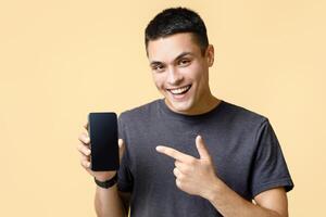 Work online, sale, modern devices and advertising of mobile application. Happy young man points finger at smartphone with empty screen, isolated on light sand background, studio shot photo