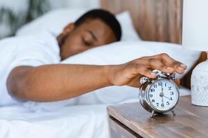 Morning Awakening. Sleepy Black Man Turning Off Alarm Clock Waking Up Early Lying In Bed In Bedroom At Home. Time To Wake-Up And Get Up Concept. Shallow Depth photo