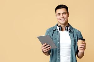 Work and break, coffee and gadgets. Cheerful young man in casual, with modern headphones, uses digital tablet and hold cup of coffee, isolated on beige background, studio shot, free space photo