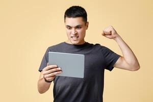 Victory online, gesture of luck and success in game. Happy excited young man in brown t-shirt looks at digital tablet and rejoices to win, isolated on beige background, studio shot, free space photo