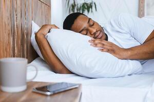 Oversleeping Concept. African American Guy Sleeping Through Alarm Clock In Morning Lying In Bed At Home, Focus On Smartphone. Rest, Nap And Recreation. Man Resting Not Hearing Alarm-Clock On Phone photo