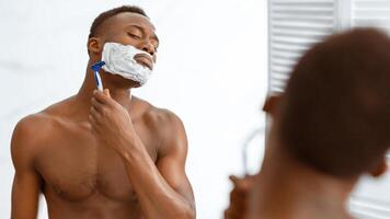Shirtless African American Man Shaving Face With Shaver Razor And Shave Foam Standing Near Mirror In Modern Bathroom At Home. Male Facial Skincare And Morning Routine. Selective Focus, Panorama photo