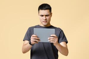 Strange , shocking information, weird message. Surprised and upset young man in brown t-shirt looks in digital tablet and reads news, isolated on beige background, studio shot, free space photo