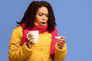 Coronavirus, Covid-19 Symptom. Sick Black Lady Having Fever, Looking At Thermometer In Shock, Measuring Temperature Standing On Purple Background, Wearing Winter Jacket. Studio Shot photo