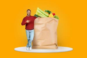 A customer stands next to a large paper bag filled with fresh groceries. The vibrant backdrop emphasizes the act of shopping in today's isolated, delivery-focused environment. photo