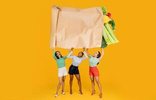 Three women joyfully raise a large paper bag overflowing with fresh groceries. They are dressed in casual clothing against a bright yellow background, reflecting the excitement of shopping. photo