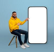 Positive arab guy pointing at big smartphone with white screen while sitting on chair over blue background. Man demonstrating free space for your app or website design, mockup photo