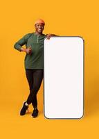 Happy Black Hipster Guy Leaning At Big Smartphone With Blank White Screen And Gesturing Thumb Up, Cheerful African Man Recommending New App Or Website, Standing On Yellow Background, Mockup Image photo