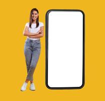 Full body length of smiling casual lady standing with folded arms near giant mobile phone, empty white screen template, orange studio background. Mock up for app or website, free space for advertising photo