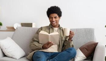 Positive black teenager reading book and drinking coffee while sitting on sofa at home. Smiling African American teen guy studying remotely from home, making assignment, looking through textbook photo