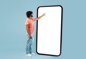 Stylish curly young hindu guy in casual outift touching huge smartphone white empty screen over blue studio background, checking newest mobile app, panorama with copy space, mockup photo