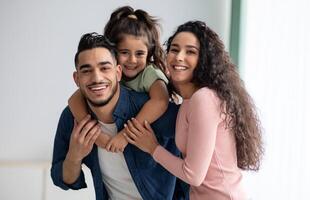 retrato de contento joven Arábica familia de Tres con pequeño hija. medio oriental mamá, papá y niño teniendo divertido mientras posando a hogar, padres disfrutando gasto hora con su niño, gratis espacio foto