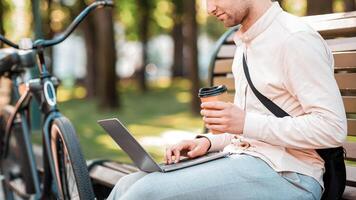 Eco transport and work in city. Young man with bag working on laptop and drinking coffee on bench in park, side view, copy space photo